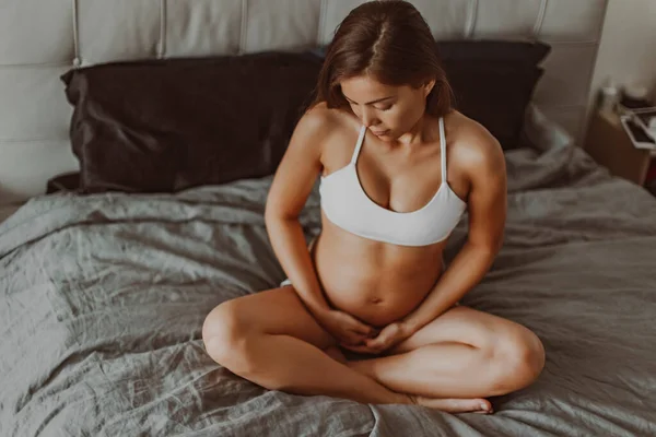 Pregnant woman sitting in bed during pregnancy thinking about her baby and motherhood. Happy multiracial asian model in first trimester showing belly and baby bump in pajamas. Maternity concept.