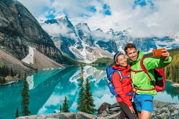 莫兰湖情侣游客使用电话在加拿大旅行远足时自拍自拍 年轻的徒步旅行者在班夫度假时打电话很开心 — 图库照片