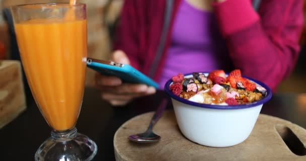 携帯電話を使ったスムージーでカフェで朝の朝食を食べるアサイーボウルの女性 果実とオートミールとの減量のためのフルーツスムージー健康的な食事療法の閉鎖 有機生ビーガン健康食品 — ストック動画