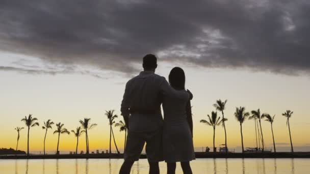 ロマンチックな夕日の幸せなカップルは 新婚旅行の休暇の休暇を受け入れるためにお互いの周りの水の景色を見て受け入れるビーチで夏の休暇の日差しを楽しんでいます レッド エピック — ストック動画