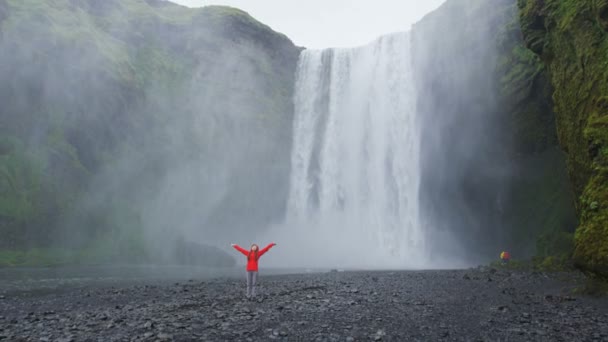一个快乐的女人通过Skogafoss瀑布在冰岛宁静和自由的户外 女孩参观冰岛自然的旅游景点和地标 红色Epic慢速运动 — 图库视频影像