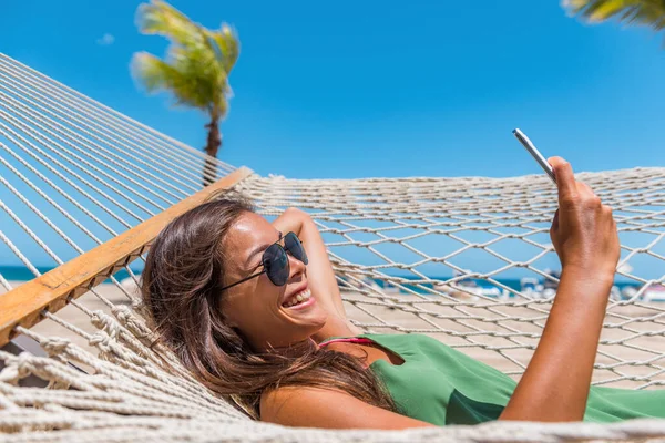 電話アプリを使用して幸せなビーチの女性熱帯の休暇にリラックスハンモックに横たわってテキストメッセージを読むSms 若いですカジュアル可愛いです女の子横たわっ屋外スイングベッド楽しむ日光浴使用して携帯電話 — ストック写真