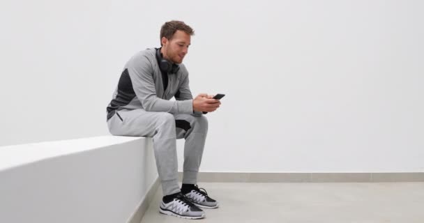 锻炼前戴耳机坐在手机应用程序上的人 健康的生活方式运动运动员在户外跑步休息时使用智能手机 穿着运动裤的男人 — 图库视频影像