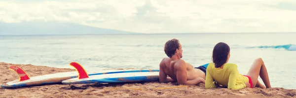 在夏威夷海滩冲浪后 情侣们放松了一下 夏威夷的一个冲浪班结束后 有两个人在日落时分躺在沙滩上 旁边的冲浪板上 观看全景 — 图库照片