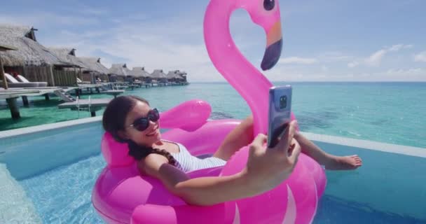 电话度假旅行妇女在充气的火烈鸟浮动床垫上使用手机在游泳池里自拍 女孩放松日光浴 享受比基尼度假度假池 — 图库视频影像