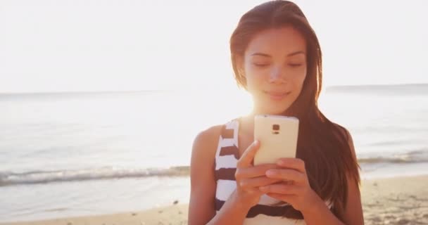 スマートフォンを使ってビーチで美しい若い女性 女の子は携帯電話でメールしてる 多人種間のアジア系中国人 白人の若い女性は 太陽のフレアと外の携帯電話を使用して笑顔 — ストック動画