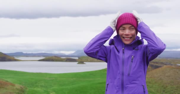 ハードシェルのジャケットと手袋を身に着けてニット帽を調整する女性は 古典的なアイスランドの天気でハイキングで頑丈なアイスランドの自然景観を楽しんで笑っています 多人種の人々 — ストック動画