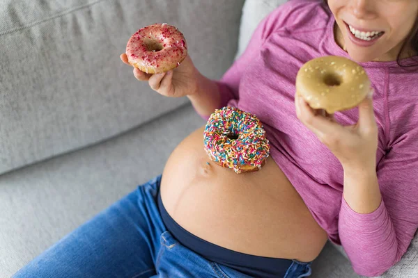 Pregnant woman eating sweet donuts on belly. Cravings of desserts and unhealthy food during pregnancy, Funny top view of cakes on baby bump for gestational diabetes or food cravings during pregnancy.