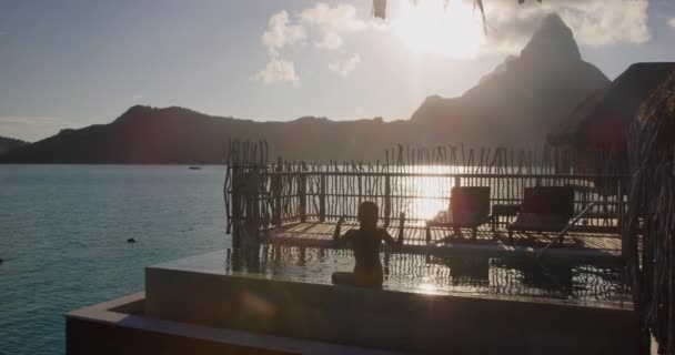 ホテルの高級リゾートハイエンド豊かな人々のライフスタイル女性観光客はボラボラ タヒチ フランス領ポリネシアの水上バンガロースイートのプライベートバルコニーテラスでインフィニティプールで夕日を見ながらリラックス — ストック動画