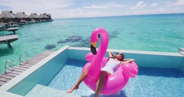 フラミンゴフロート夏休みスイミングプールで膨脹可能なピンクのプールのおもちゃのマットレスのビキニの女性 リゾートプールで旅行の休日を楽しむエレガントな女性のリラックス日光浴 ラグジュアリーライフスタイル — ストック動画