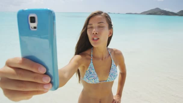 ビーチでスマートフォンを使用している携帯電話の女の子は 夏の旅行休暇中に自撮り写真を撮るか ビデオチャットをしている間にビーチ休暇で楽しいキスを吹いています セクシー若いですビキニ女性ポルノのカメラ — ストック動画