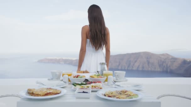 女性が歩いてテラスで朝食テーブル 海と山のリゾートで提供されるおいしい料理 カルデラを眺めながらサントリーニ島の休暇旅行休暇の観光客 — ストック動画