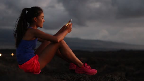 女人在晚上跑步后用智能手机看健身追踪应用程序 跑步运动和健身的女跑步者在外面放松地看着智能手机屏幕 健康的生活方式 美国夏威夷 — 图库视频影像