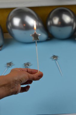 A hand holding a silver star-shaped candle against a light blue background with silver balloons and matching candles in the frame. Perfect for celebrations, parties, or festive themes with a modern touch. clipart