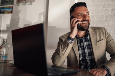 Akıllı telefon kullanan yakışıklı bir iş adamı ve bir kafede kahve içerken dizüstü bilgisayarı üzerinde çalışıyor.