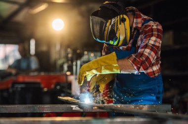 Koyu renk saçlı bir adam projesi için metal bir yapıyı kaynak yaparken bir atölyede sıkı çalışıyor. Kaynak maskesini takmış, güneşlik takmış, gözlerini parlak kıvılcımlardan koruyor..