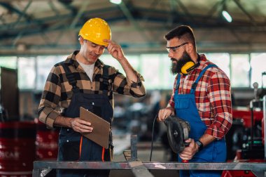 Metal atölyesinde iş arkadaşları ürünleri öğütmeye hazırlanıyor. Sarı başlıklı usta, iş arkadaşının gerekli ölçümleri alıp almadığını kontrol ediyor. Boşluğu kopyala.