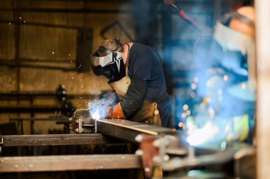 Çalışkan bir adam, etrafındaki sarı kıvılcımlar uçuşurken metal bir plakayı titizlikle ve konsantrasyonla kaynaklıyor. Zor ve tehlikeli bir çevrede çalışmaya adanmışlık. Boşluğu kopyala.