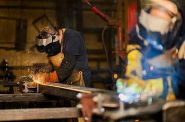 Kafkasyalı bir adam atölyede metal bir plaka kaynak yaparken iş yüzeyinde altın kıvılcımlar uçuşuyor. Koruma için güvenlik eldiveni ve kaynak maskesi takıyordu. Tehlikeli bir iş ortamı. Boşluğu kopyala.
