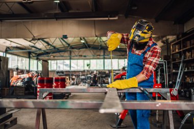 İş tulumu ve uzun kollu ekose gömlek giyen sakallı, koyu renk saçlı bir adam bir mezura ile kesin ölçüler alıyor. Güvenlik eldivenleriyle metal plakayı tutuyor ve ona bakıyor. Boşluğu kopyala..