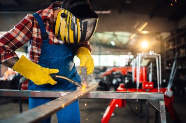 Tehlikeli bir iş ortamında güvenlik ekipmanı takıp uçan kıvılcımlara bakarken metal kaynak yapan işçiler. Çalışma alanının bulanık arkaplanı ve araç gereçlerle raflar. Boşluğu kopyala.