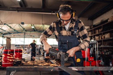 Kulaklık ve gözlüklü bir işçiye odaklan. Bir yandan kıvılcımlar uçuşurken diğer yandan metal bir yapıyı öğütüyor. Metal fabrikasının kapalı arka planı bulanık. Boşluğu kopyala.