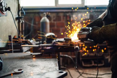 Güçlü bir öğütücüyle çalışırken koruyucu eldiven takan bir zanaatkârın ellerine odaklan. Atölyenin her yerinde altın kıvılcımlar uçuşurken metal bir levhayı öğütüyordu. Boşluğu kopyala.