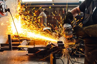 Güçlü bir öğütücüyle çalışırken koruyucu eldiven takan bir zanaatkârın ellerine odaklan. Atölyenin her yerinde altın kıvılcımlar uçuşurken metal bir levhayı öğütüyordu. Boşluğu kopyala.