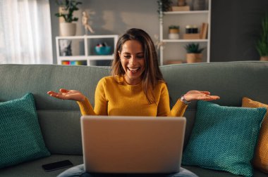 Evdeki kanepede otururken ellerini açıp gülümseyen mutlu, beyaz bir kadın. Sosyal ağda mutlu kadın alışverişleri ya da sohbet, serbest çalışan bir proje üzerinde çalışan.