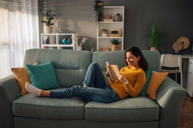 Evde boş zamanlarında kanepede uzanmış kitap okuyan genç bir kadının portresi. Oturma odasındaki kanepede dinlenen güzel bir kadın. Boşluğu kopyala.