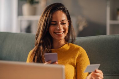 Mutlu kadın evde oturuyor, gülümsüyor ve telefonla online alışveriş yapmak için kredi kartı kullanıyor. Kart detaylarını okuyor ve telefonda yazıyor. Gülümseyen kadın online alımını doğruluyor..