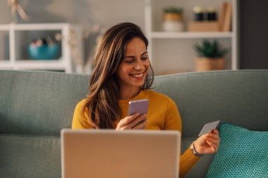 Neşeli bir kadın evdeki koltukta oturuyor ve çevrimiçi alışveriş için kredi kartı, telefon ve dizüstü bilgisayar kullanıyor. İnternetten alışveriş ve internetten sipariş. Teknoloji kullanan kadın portresi.