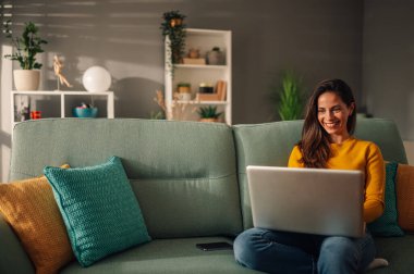 Neşeli bir kadın kanepede rahat evinde oturuyor ve ona gülümserken dizüstü bilgisayar kullanıyor. Mutlu genç bir kadın interneti ve interneti aramak için teknolojileri kullanıyor. İç mimari