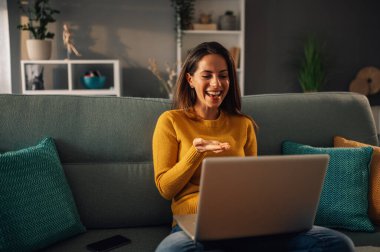 Neşeli bir kadın rahat dairesinde bir kanepede oturuyor ve dizüstü bilgisayarında bir video görüşmesi yapıyor. Bir kadın internette konferans görüşmesi yaparken dizüstü bilgisayara gülümsüyor..