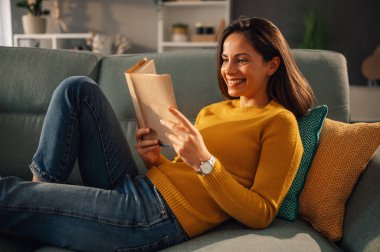 Genç ve mutlu bir kadının portresi kanepede uzanıyor ve rahat evinde kitap okuyor. Mutlu bir kadın bir roman okuyor ve kitaba gülümserken rahat dairesinde dinleniyor..