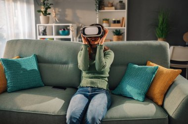 Genç bir kadın evdeki kanepede oturuyor ve sanal gerçeklik deneyimi yaşarken kafasında kulaklık taşıyor. Mutlu bir kadın evinde teknoloji kullanarak eğleniyor..