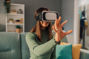 Genç bir kadın kafasında sanal gerçeklik kulaklığıyla evde oturuyor ve elleriyle uzanıyor. Bir kadın sanal gerçekliği evinde yaşıyor. Teknoloji harikası eğlence..