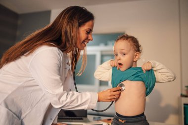 Kadın bir pediatrist, doktorun muayenehanesinde dururken steteskop kullanıyor ve küçük çocuğun akciğerlerini inceliyor. Hasta küçük bir çocuk muayene için doktor ofisinde oturuyor ve onu ziyaret ediyor..
