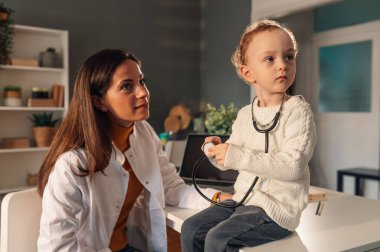 Düşünceli küçük bir çocuk doktorun muayenehanesinde bir çocuk doktorunu ziyaret ediyor. Genç bir hasta, doktorunun muayenehanesinde kadın pediyatristiyle otururken steteskopla oynuyor..