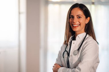 Kendine güvenen genç bir doktor iş yerinde kollarını kavuşturup kameraya gülümsüyor. İnsanlık, tıbbi yardım ve sağlık sigortası kavramı. Kameraya gülümseyen dost canlısı bir doktor.