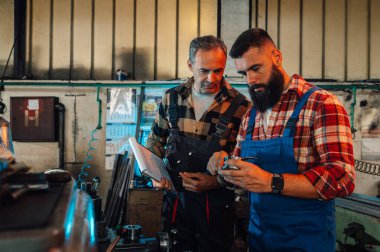 Bir atölyedeki metal parçasının ölçüleri hakkında danışmanlık yapan iki metalürji uzmanı. Tulum pantolon ve ekose gömlek giyiyorlar. Sakallı genç işçinin elinde bir kalibre var..