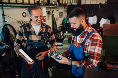 Sakallı genç bir makinist son çalışmalarını kıdemli bir mühendise gösteriyor. Makinist elinde bir dosya tutuyor, makinist elinde iki rulo kesiği tutuyor. Tulum ve gömlek giyiyorlar..