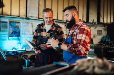 Bir atölyede metal parçasını inceleyen iki metalürji işçisi. Kıdemli mühendis bir dosya tutuyor, genç makinist vites tutuyor. Ekose gömlek ve tulum giyiyorlar. Bulanık arkaplan.