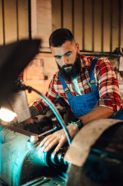 Torna makinesini çalıştıran bir makinistin resmi rulo kesim bir parça yaratmak için. Metali sıkıyor. Ön plan bulanık, fabrika ayarları, atölye çalışması. Bir işçi makineyi tamir ediyor ve ayarlıyor.