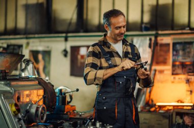 Üst düzey metal çevirici atölye fabrikasındaki torna makinesinin yanında çelik taşıyıcı ölçüyor. Arka plandaki sıcak turuncu ışık makinenin yüzeyine yansıyor. Tulum giyiyor..