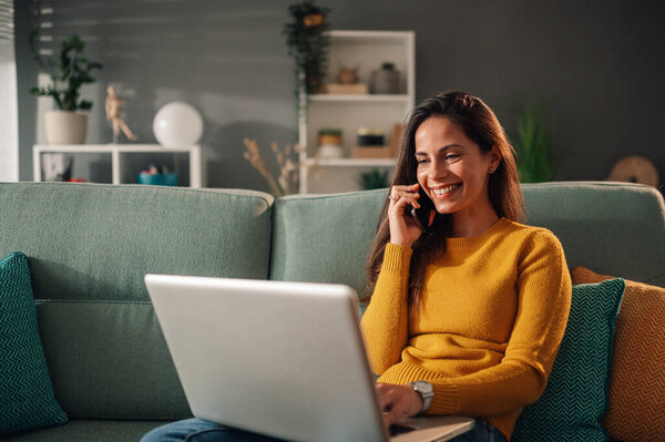 A cheerful woman is sitting on couch at cozy apartment and having a phone call while typing on a laptop. A cheerful woman is sitting at home and using technologies for online communications.