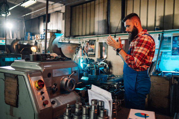 A young bearded metallurgist in his metal workshop, standing next to a lathe machine (metallurgy machine), examining an object in their hands. He has a beard and wears a shirt and overall pants.