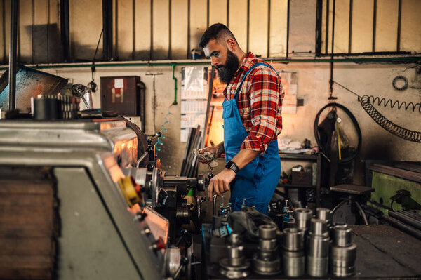 An image of a metal turner standing next to a lathe machine and a table with various metal pieces. He is operating the machine and holding a cloth. Small factory, workshop setting.Overalls and shirt.