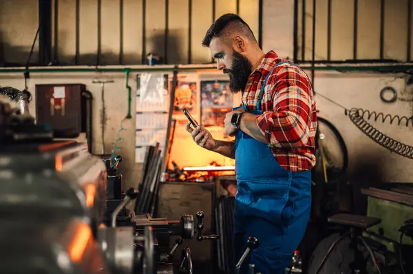 stock image A metallurgy artisan looking at the phone in front of a metalwork machine in a manufacture. He is wearing a wristwatch, red and white plaid shirt, and overall pants. Beard, tattoo. Blurred foreground.