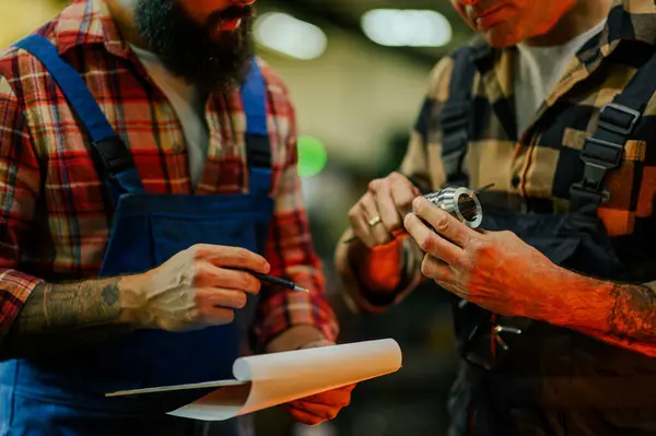 stock image Cropped picture of a mechanist and a supervisor. The mechanist is holding a machine part, measuring it with a caliper. The young man is holding a clipboard and a pen. Visible tattoos, overalls.